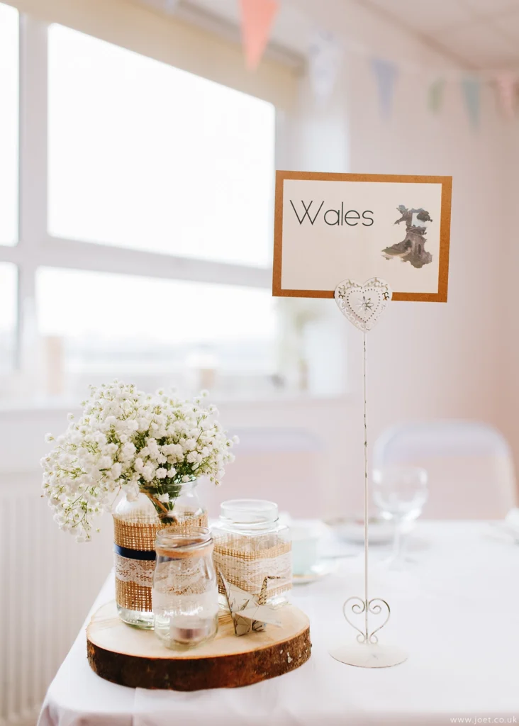 Wedding reception table decor - flowers on a log with bunting in the background. Table name reads "Wales"