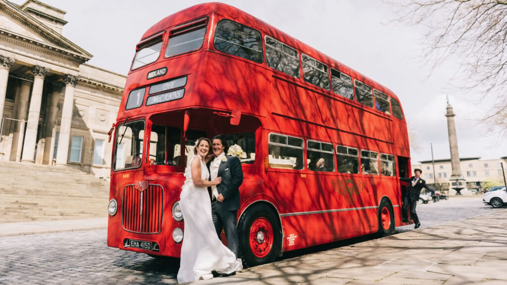 A smiling bride and groom pose joyfully in front of a vintage red double-decker bus, with a grand classical building and a monument visible in the background. This wedding photo captures a romantic, nostalgic moment on a historic British bus.